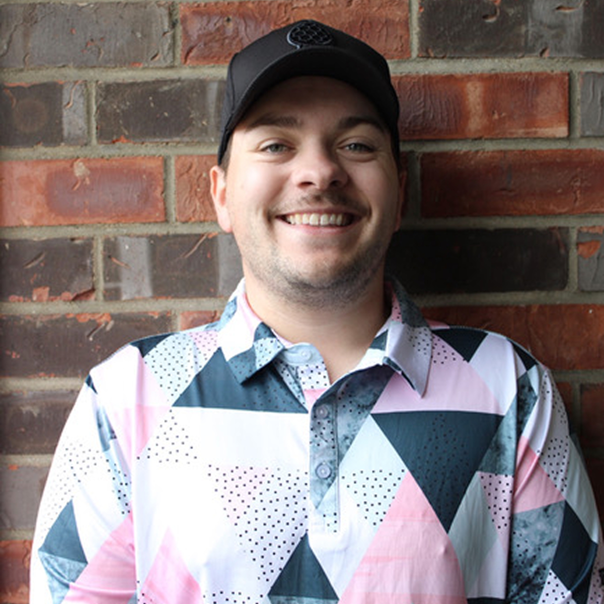 Person smiling in a geometric patterned shirt and cap, standing against a brick wall.
