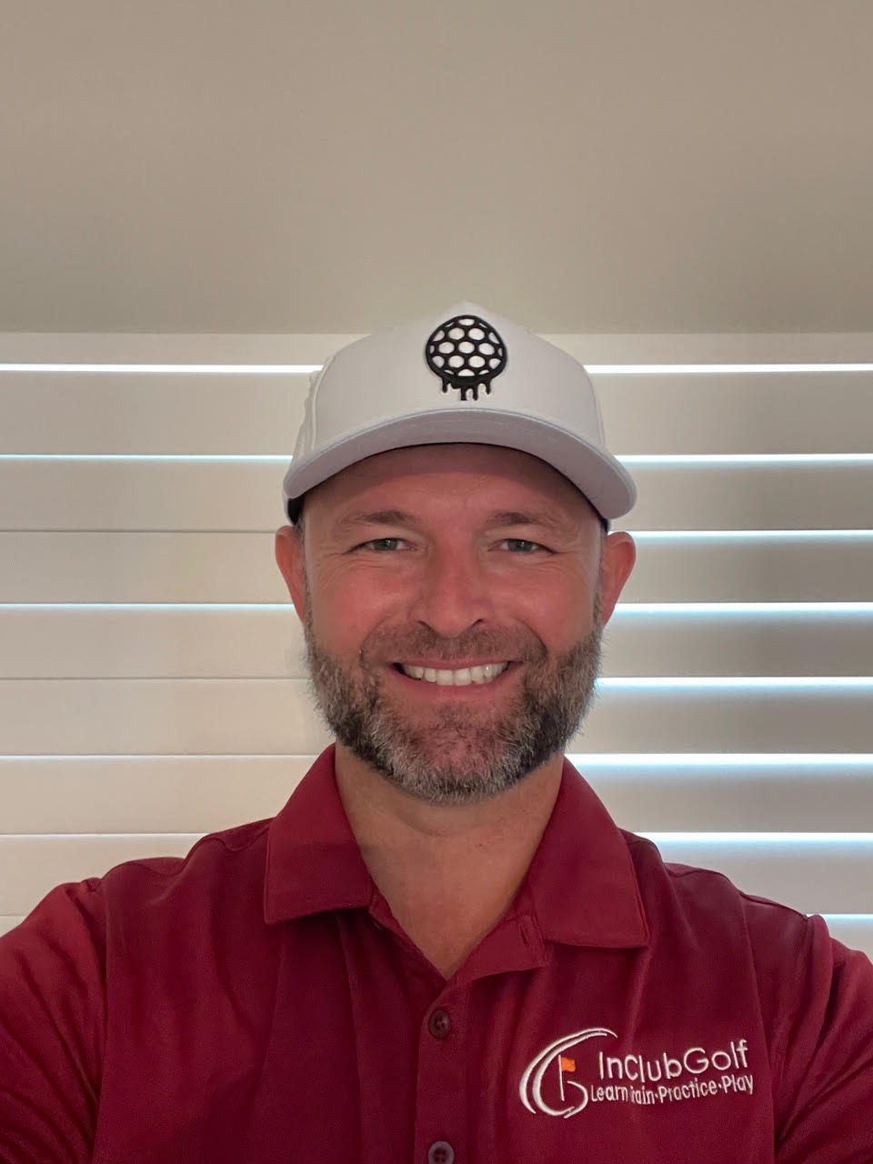 A smiling man with a beard, wearing a white hat and red polo shirt, stands before window blinds.