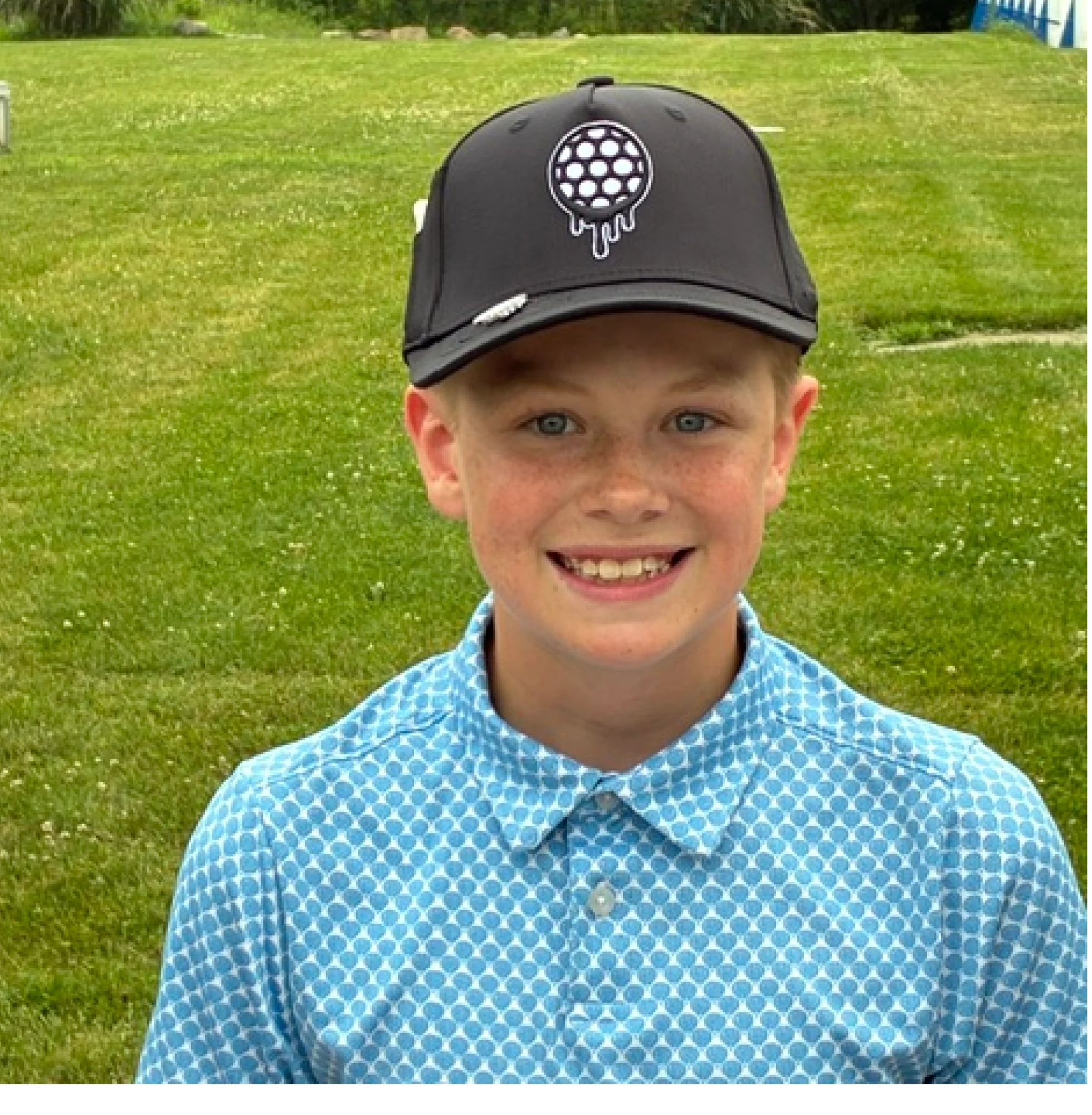 Smiling child wearing a patterned blue shirt and black cap on a grassy field.