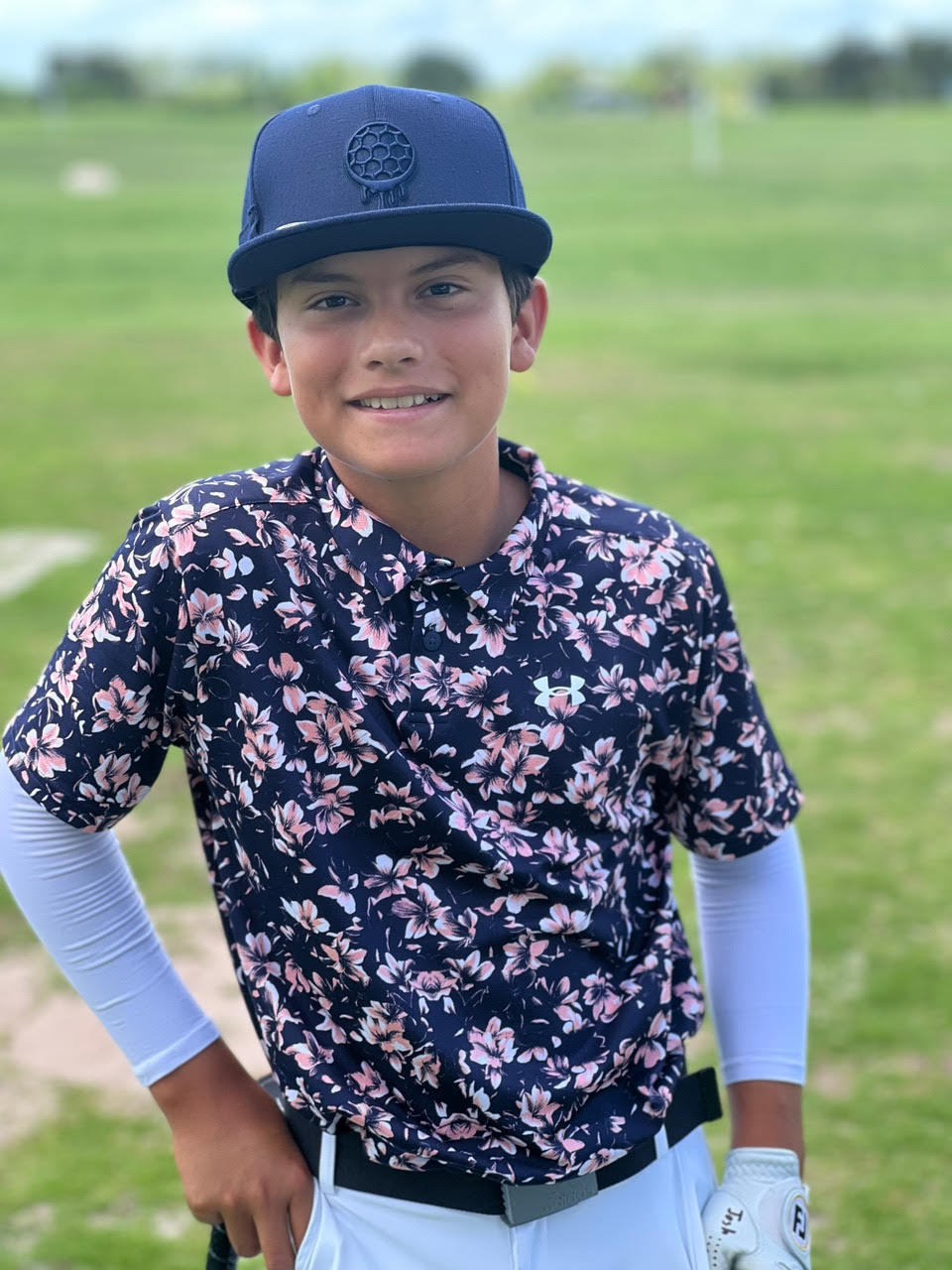 A smiling young boy in a floral shirt and a baseball cap stands on a golf course.