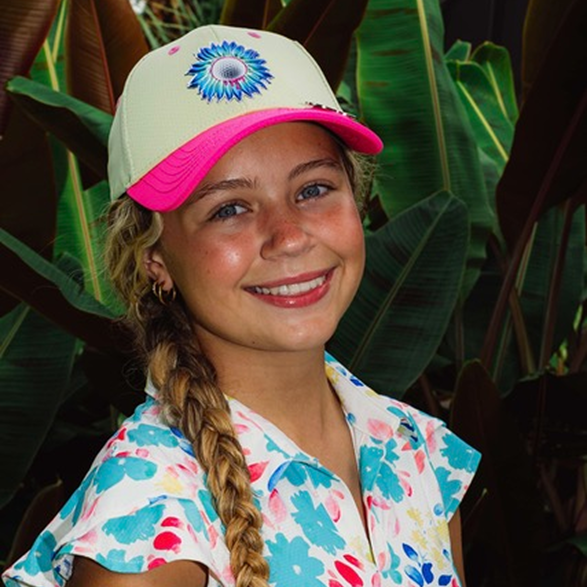 Smiling person in a floral shirt and cap, standing in front of large green leaves.