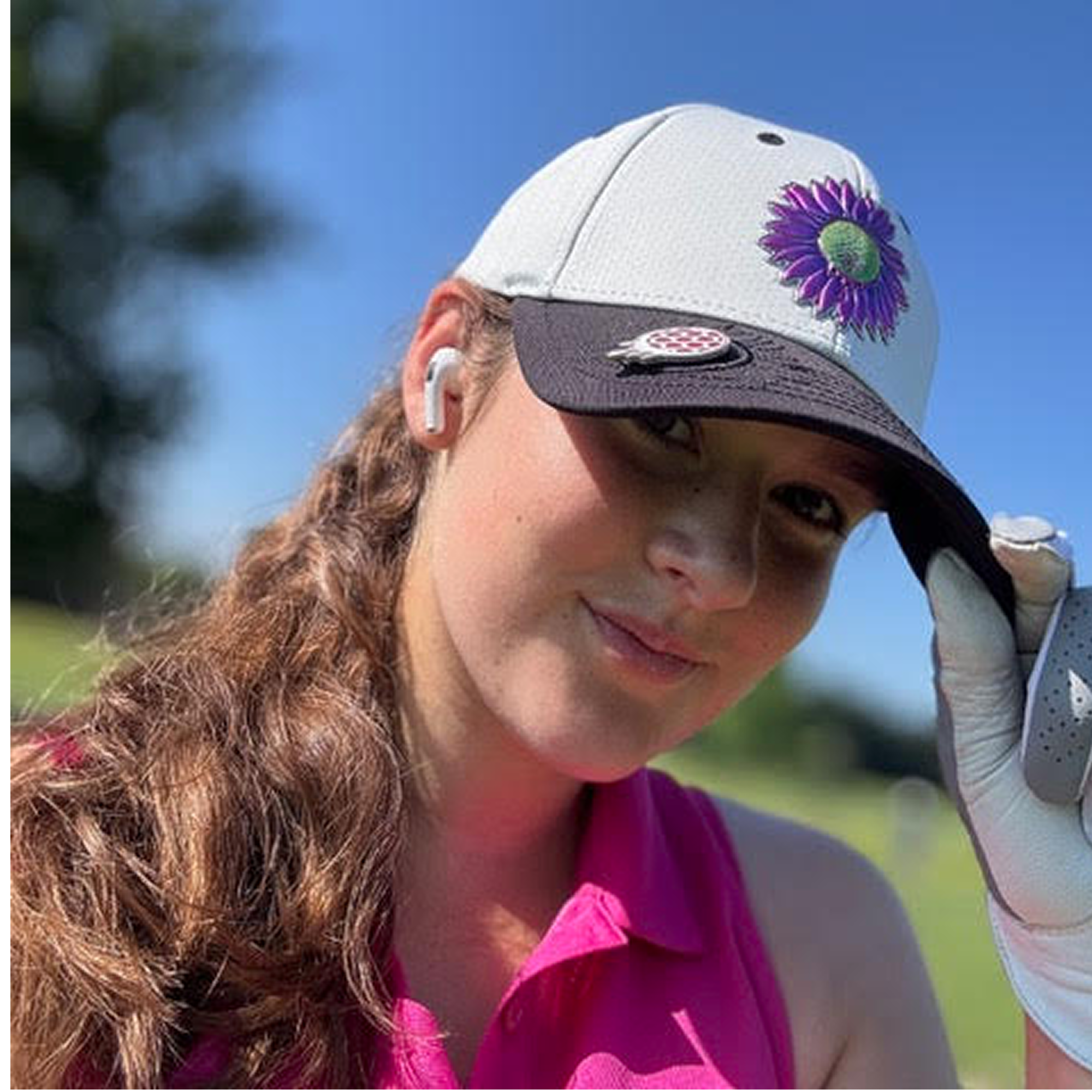 Person in a pink shirt and cap with a flower emblem, wearing earbuds outdoors.