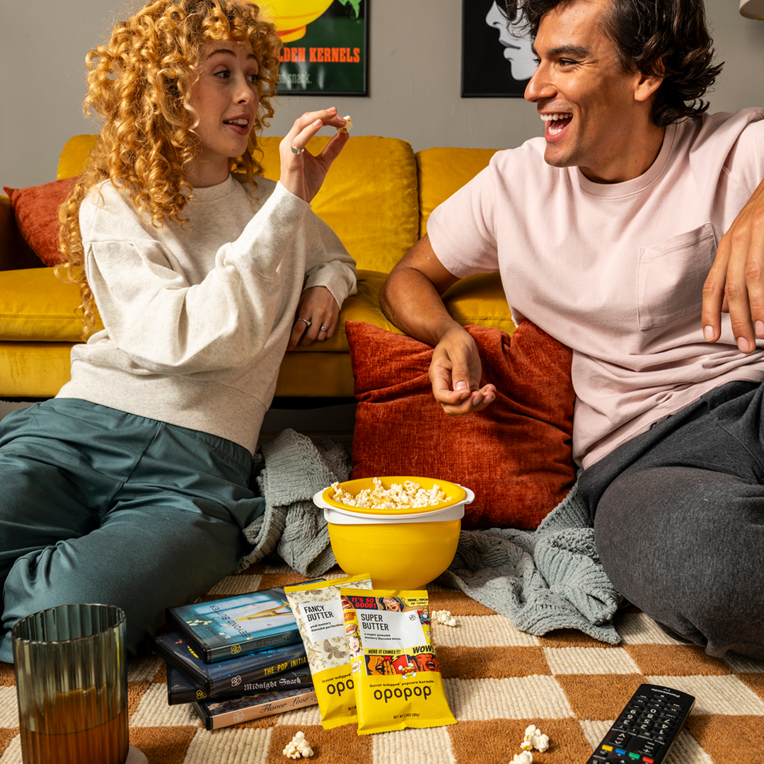 Two people sitting on a carpet with popcorn and snacks around them.