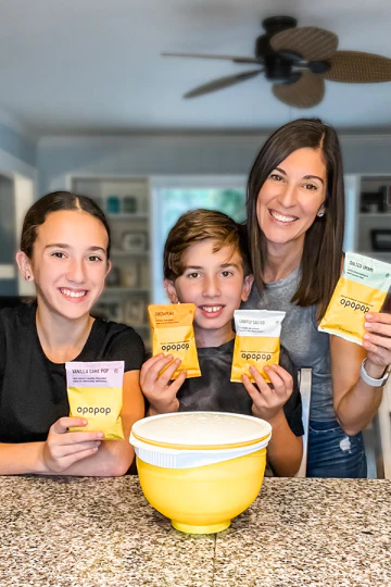 Three people holding popcorn packets in a kitchen with a yellow bowl.