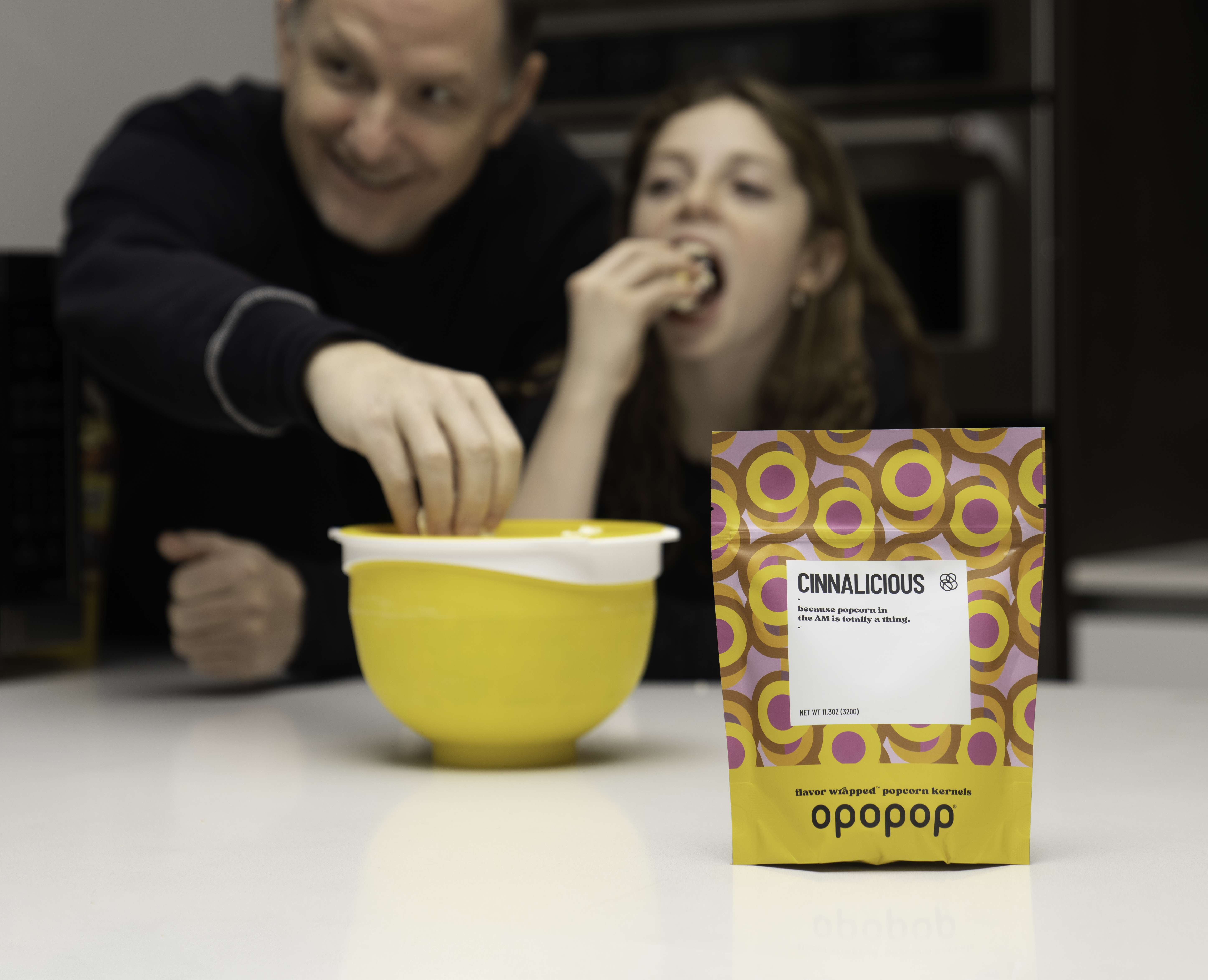 Two people eating popcorn from a yellow bowl, with a popcorn bag labeled 'Cinnalicious' in the foreground.