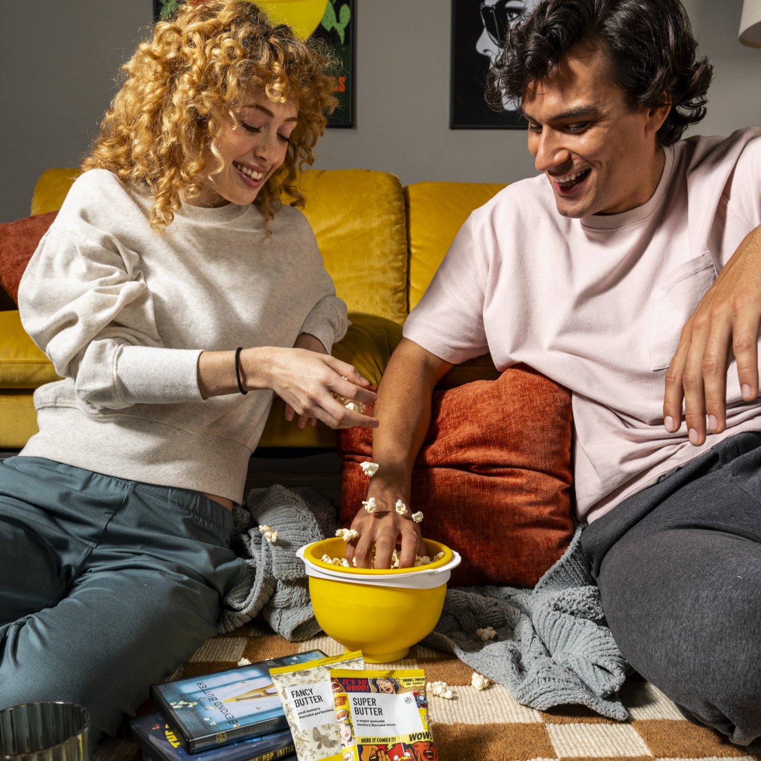 A smiling man and woman sit on the floor, playfully sharing popcorn from a large yellow bowl.