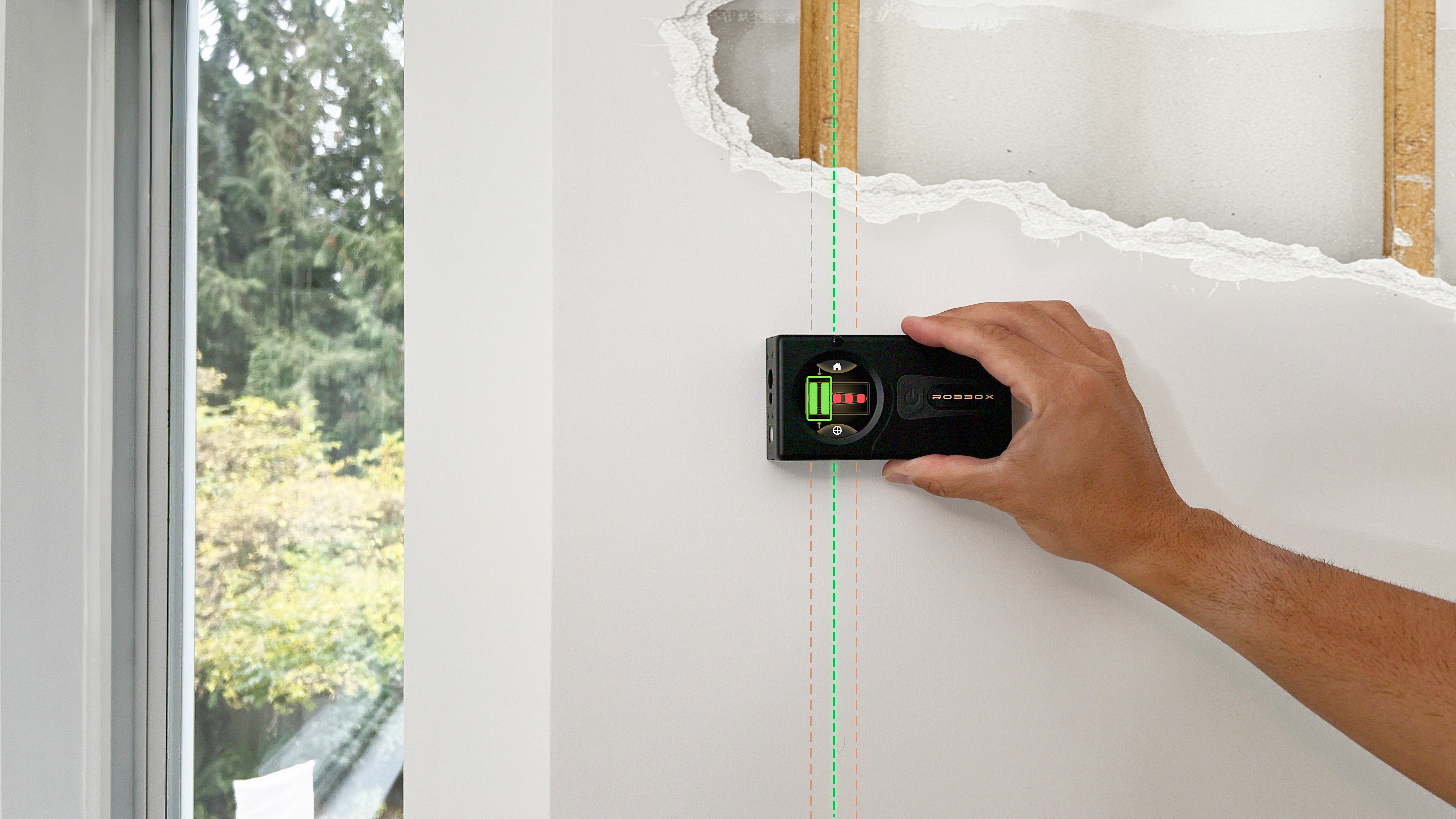 A hand using a stud finder on a white wall near a window.