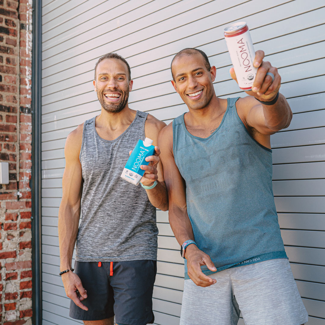 Two men in tank tops holding NOOMA drinks, smiling in front of a garage door.
