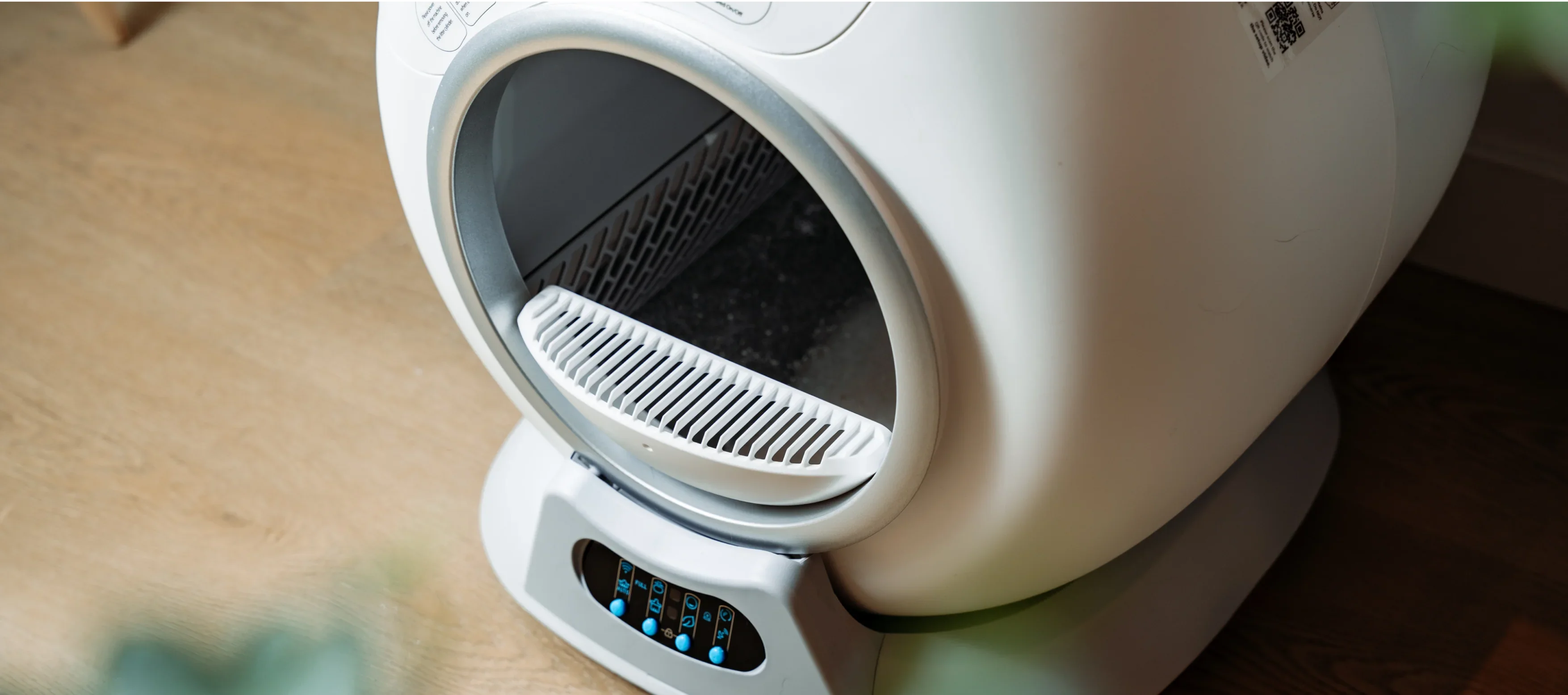 A close-up of a white, automatic self-cleaning cat litter box on a wooden floor.