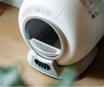 A white, modern self-cleaning cat litter box with blue lights on its control panel, on a wood floor.