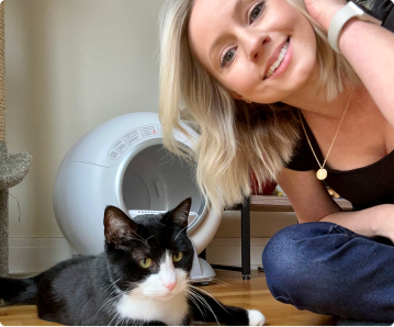 A smiling woman sitting on the floor with a black and white cat and an automatic litter box.