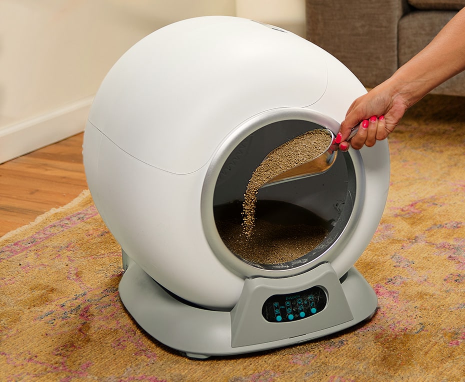 Person scooping litter into a futuristic, spherical litter box on a carpeted floor.
