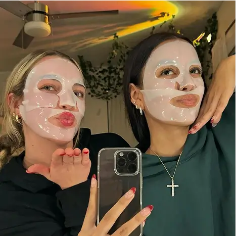 Two people wearing sheet masks pose for a selfie in a room with plants and ceiling fan.