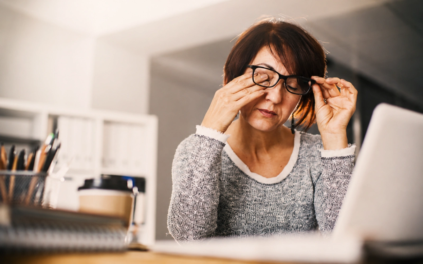 A woman wearing glasses sits at a desk in front of a laptop, rubbing her eyes.