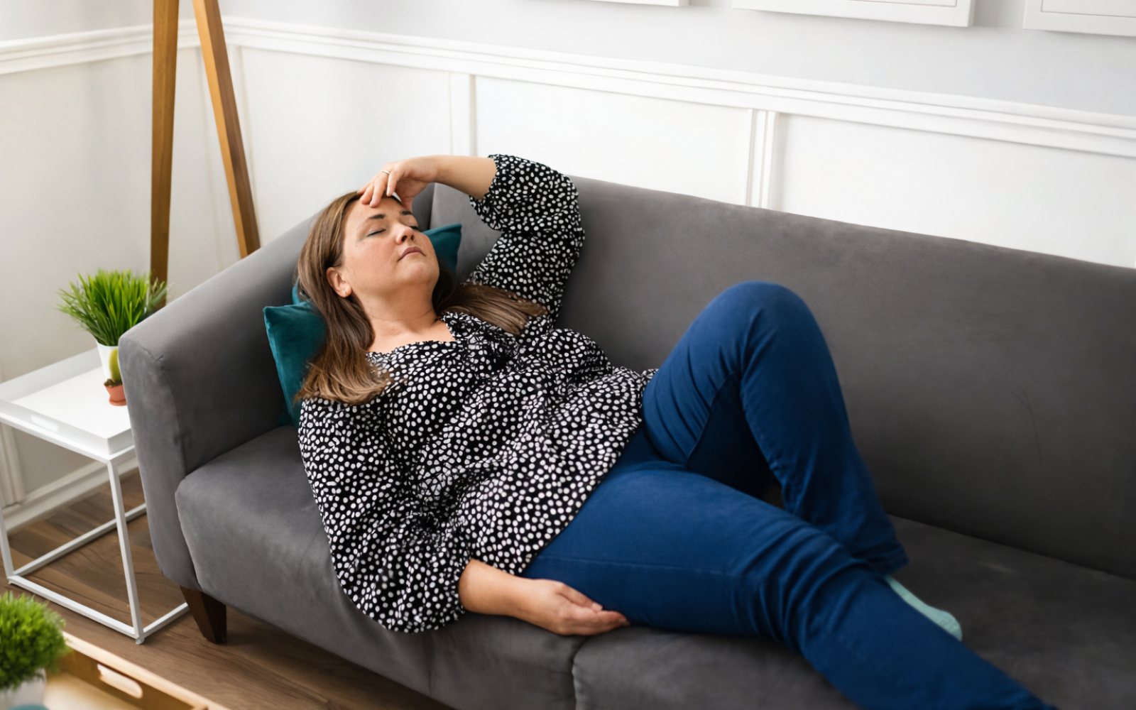 A woman lies on a grey couch with her eyes closed and a hand on her forehead.
