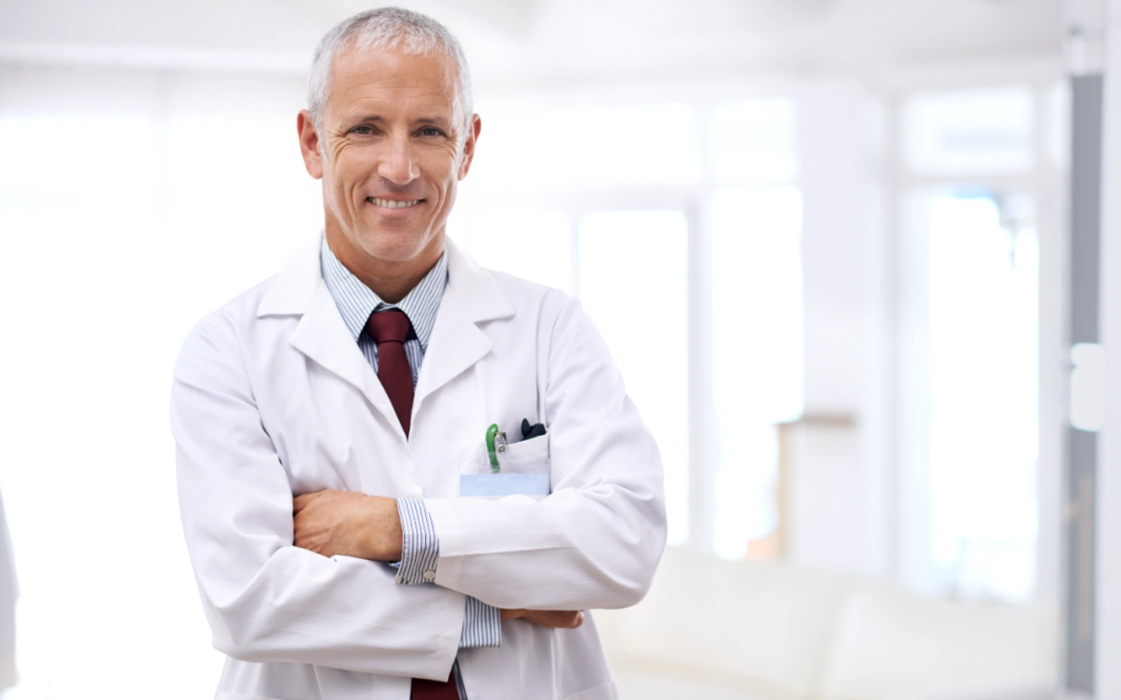 A smiling, gray-haired male doctor in a white coat stands with his arms crossed in a brightly lit room.
