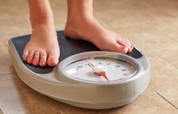 A person's bare feet with pink toenails standing on an analog bathroom scale on a tiled floor.