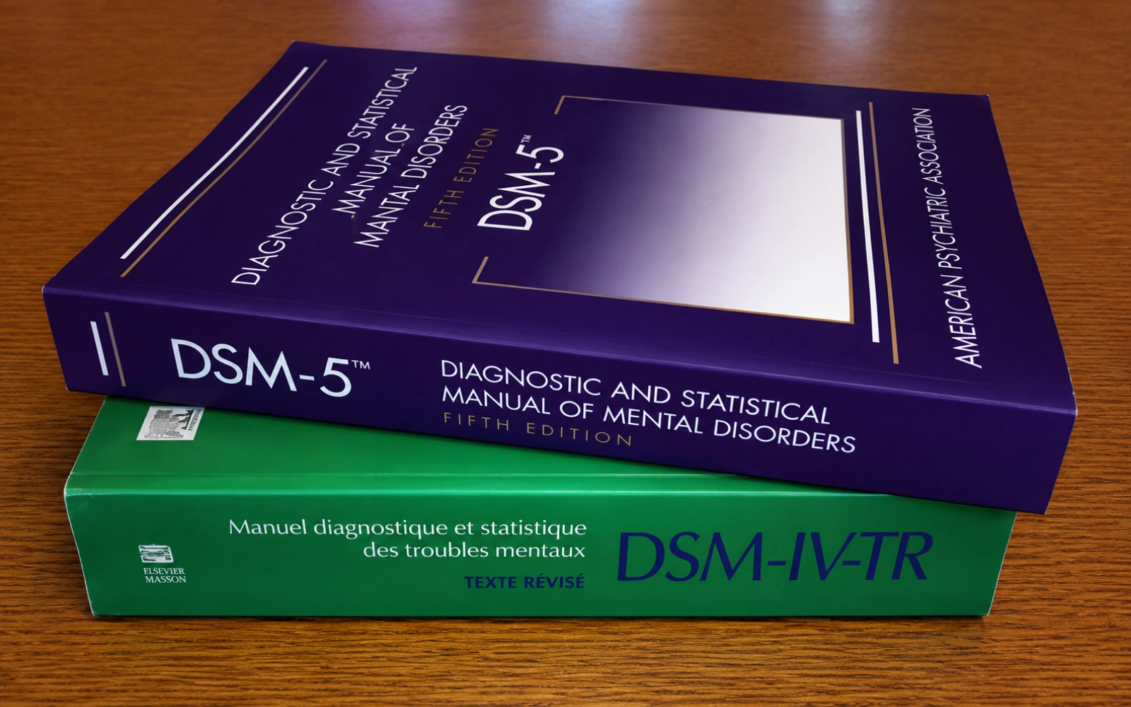 The DSM-5 and the French DSM-IV-TR books stacked on top of each other on a wooden surface.