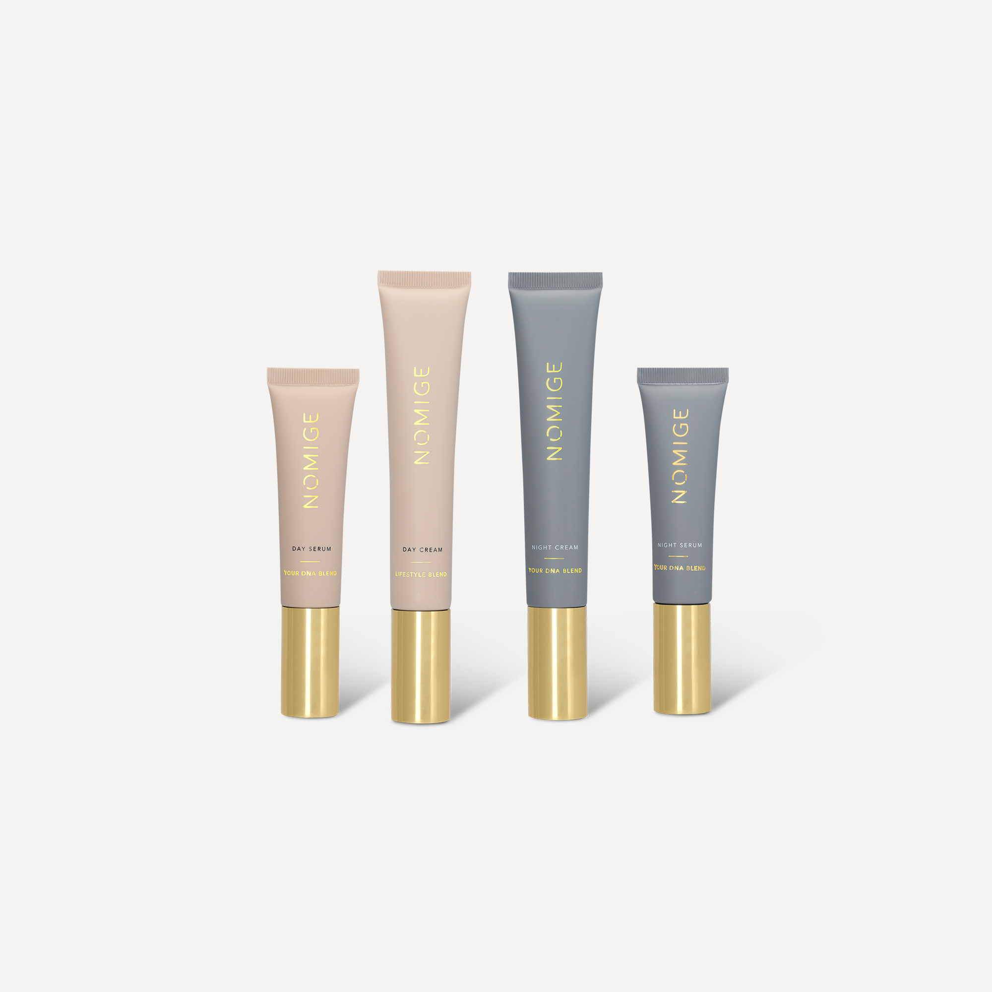 Four Nomige skincare tubes, two beige and two grey, with gold caps standing in a row.