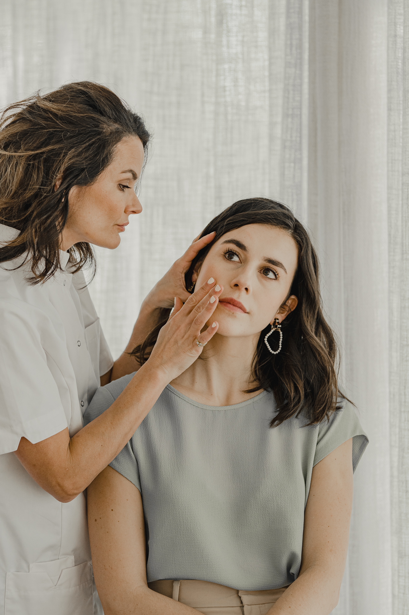A woman in a white coat examines another woman's face.