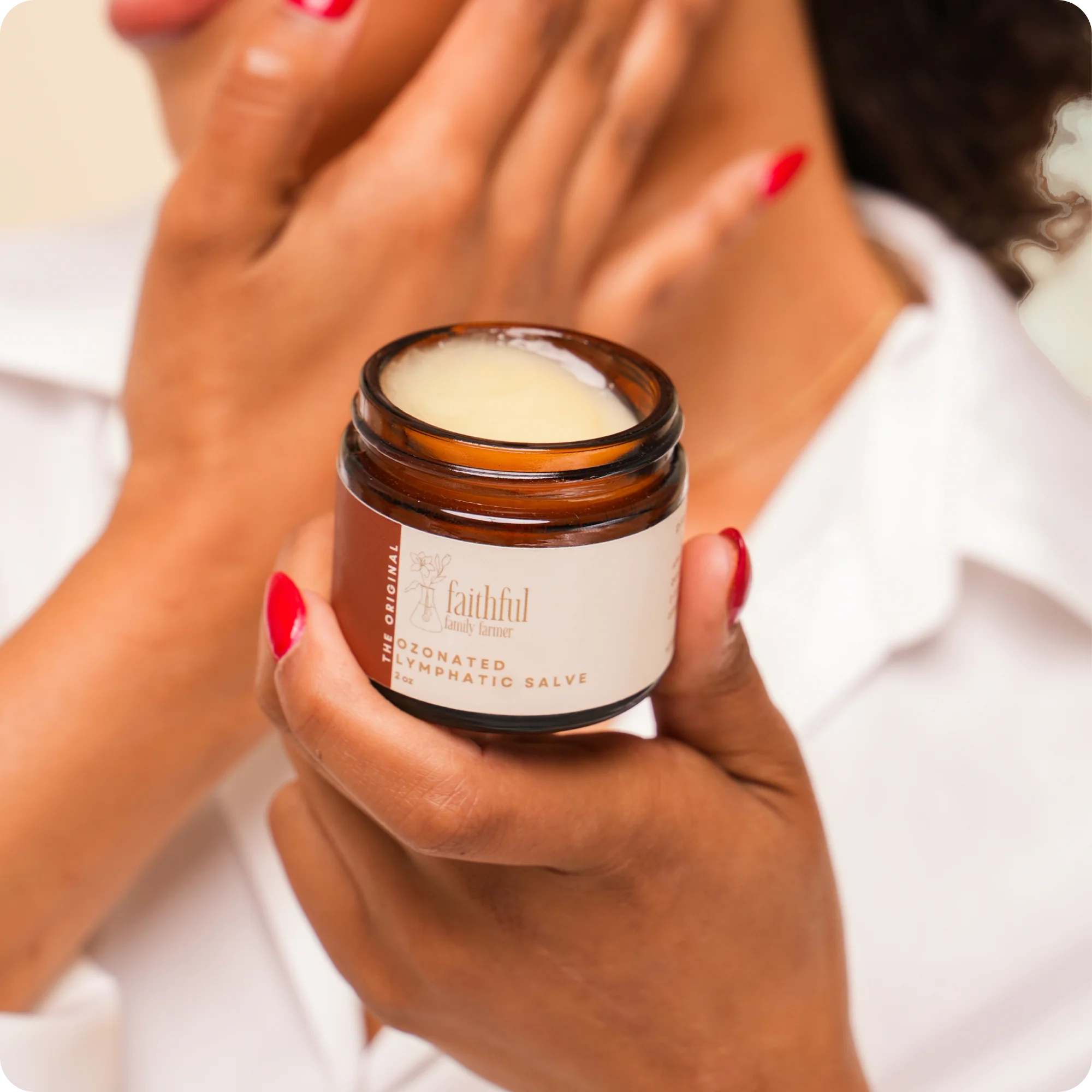 A person with red nails holds a jar of Ozonated Lymphatic Salve while touching their neck.