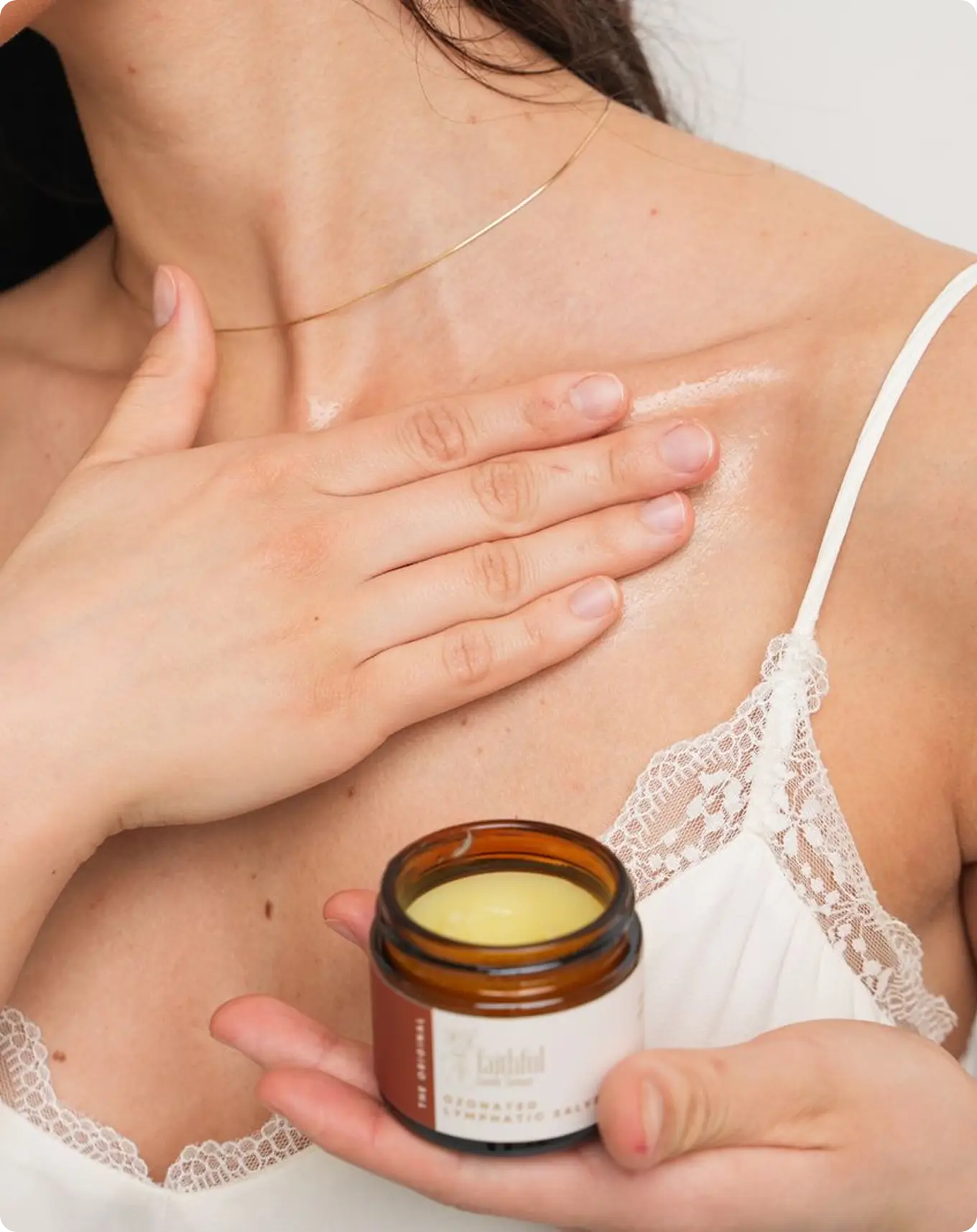 A woman in a white camisole holds a small jar while applying a balm to her collarbone.