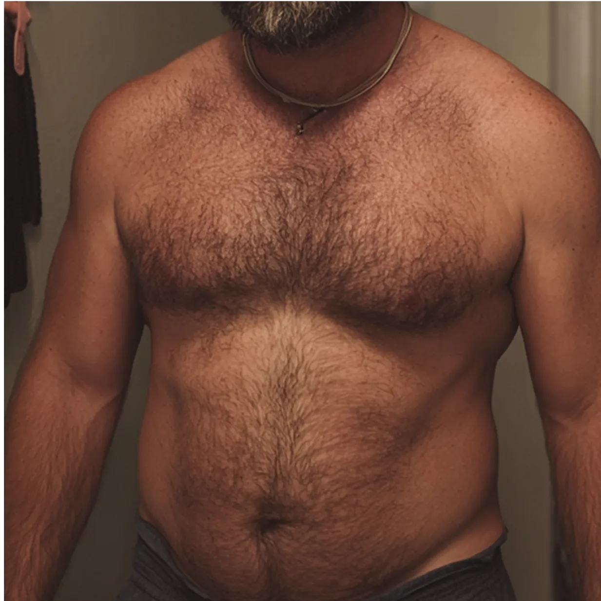 Shirtless man with a beard and a necklace in a dimly lit room.