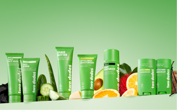 A lineup of 'two dudes' skincare products in green packaging, surrounded by fresh fruits and plants.