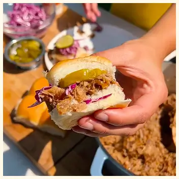 A hand holds a small pulled pork slider with red cabbage slaw and a pickle slice.