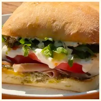 A close-up of a sandwich on a ciabatta roll with pesto, deli meat, tomato, and a white sauce.