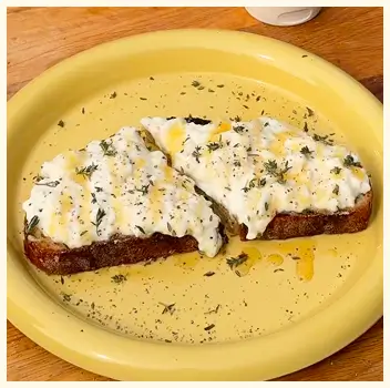 Two slices of toast with a creamy white topping, drizzled with oil and herbs on a yellow plate.