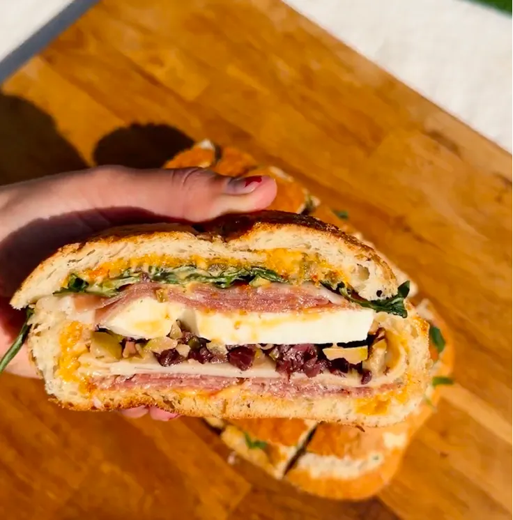 A hand holds a large sandwich cut in half, showing layers of meat, cheese, and olives.
