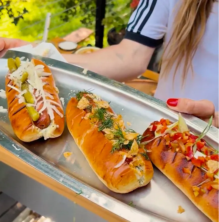 A person holds a silver platter with three elaborately-topped hot dogs.