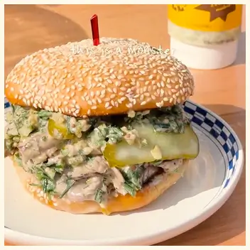 A close-up of a sandwich with a sesame seed bun, filled with a creamy salad and pickles.
