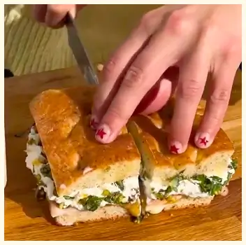 A person's hands with red stars on their nails cutting a large sandwich on a wooden board.