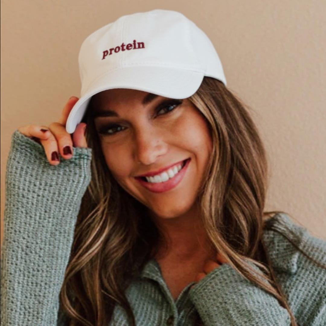 Smiling person wearing a white cap with 'protein' written on it, and a gray textured sweater.