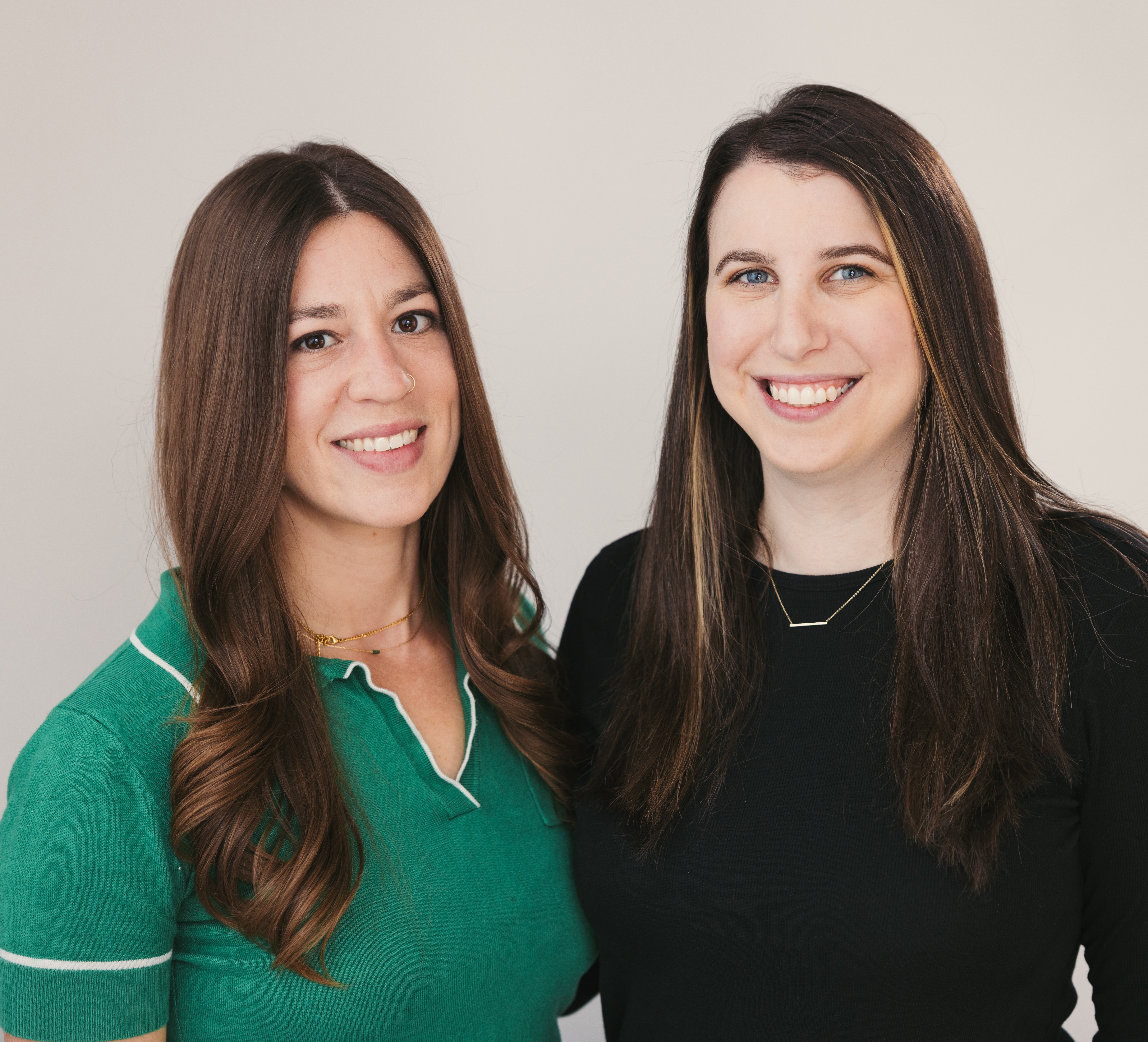 Two women smiling, one in a green shirt, the other in black, against a plain background.