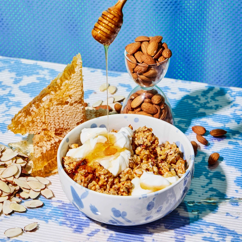 A bowl of granola and yogurt is drizzled with honey, surrounded by almonds and honeycomb.
