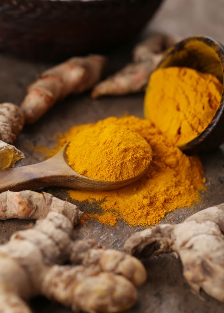 A wooden spoon with turmeric powder surrounded by turmeric roots.
