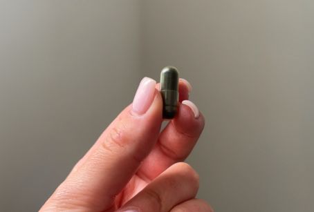 A hand holding a small green capsule against a plain background.