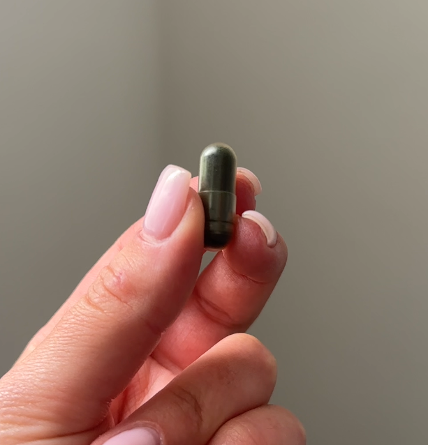 Hand holding a small green capsule against a neutral background.