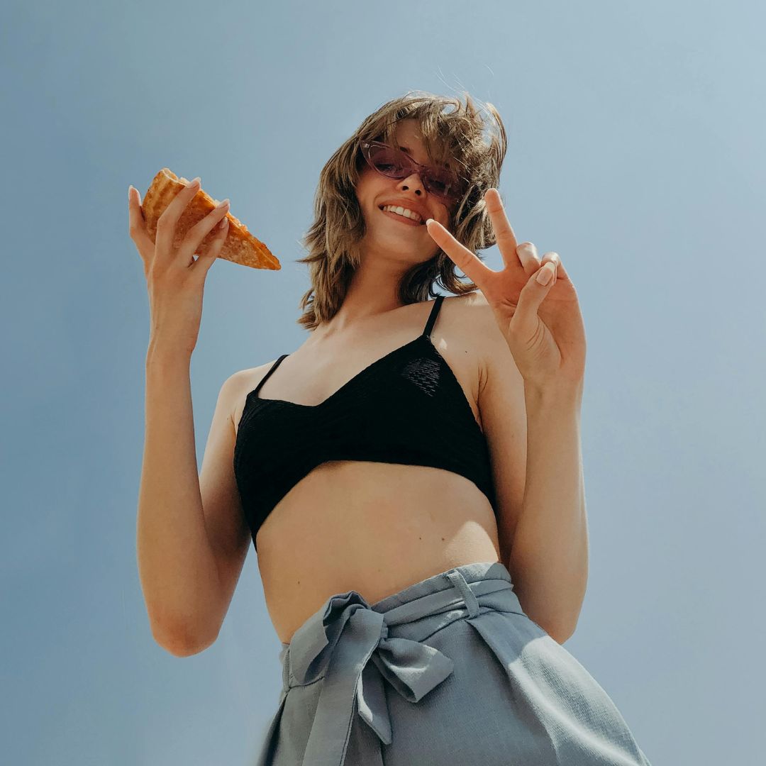 Person in sunglasses holding pizza and making a peace sign under a clear sky.