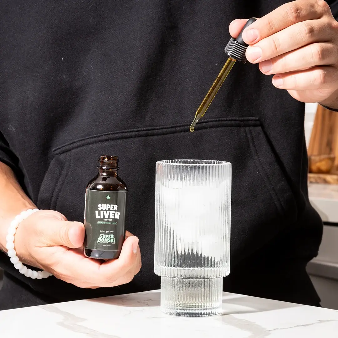 A person adds drops from a Super Liver tincture bottle into a glass of ice.