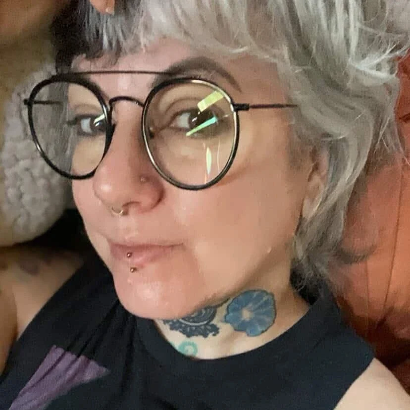 A person with short grey hair, glasses, facial piercings, and neck tattoos looks at the camera.
