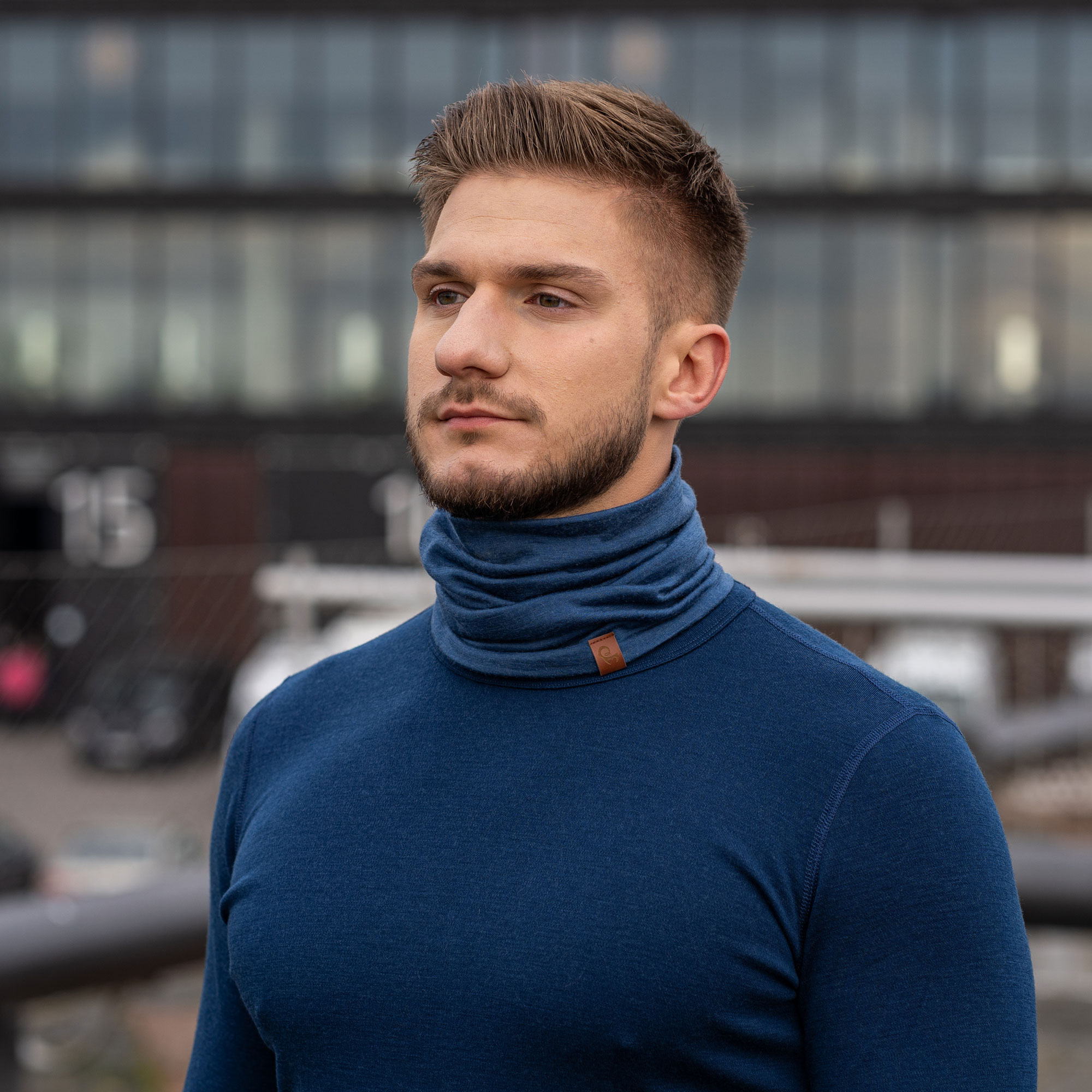A young man with a beard wearing a blue turtleneck shirt looks off to the side.