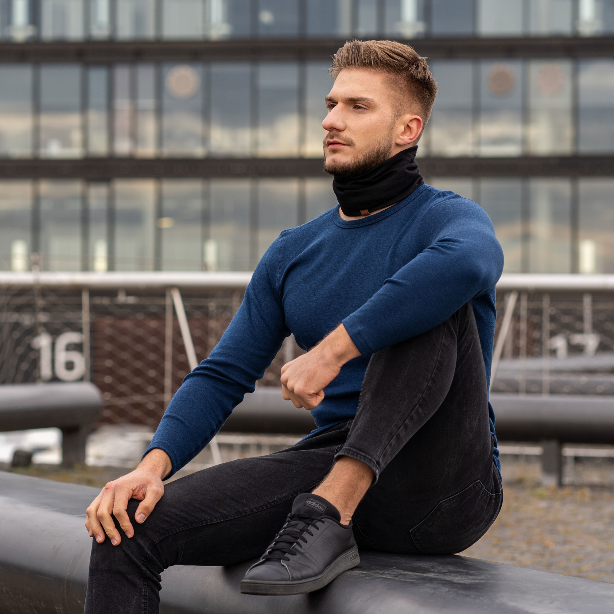 A young man in a blue long-sleeved shirt and black neck gaiter sits outside on a beam.