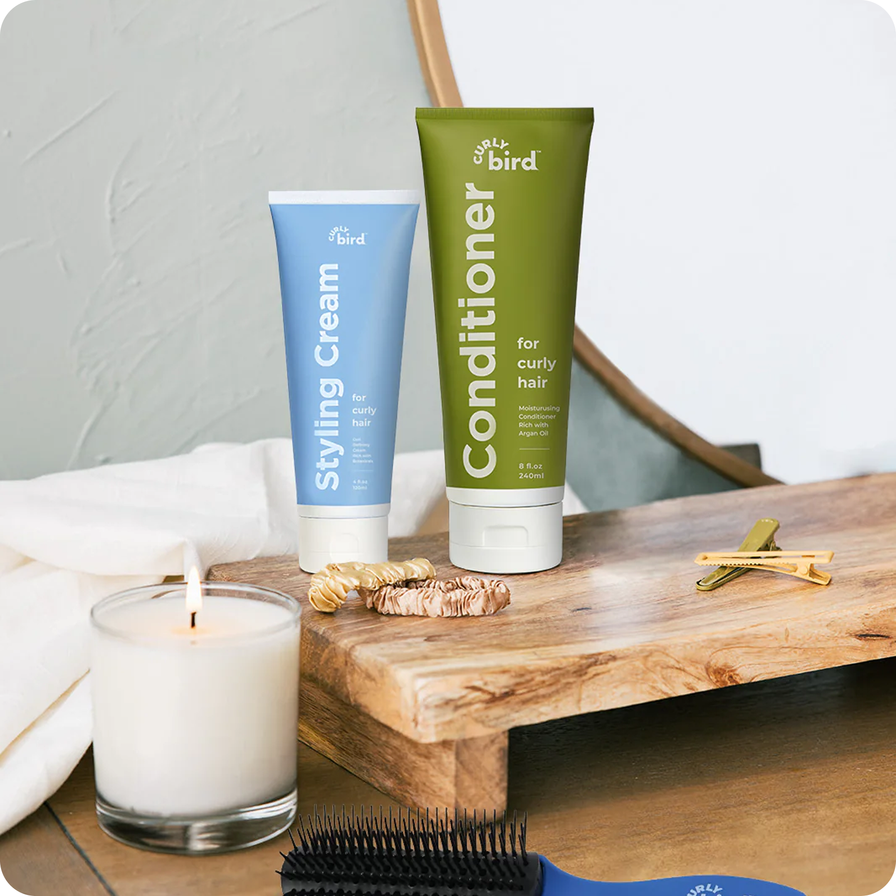 Curly Bird styling cream and conditioner on a wooden tray with a candle, brush, and hair accessories.