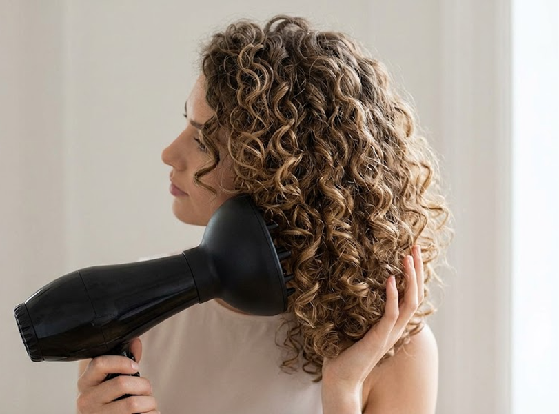 Side profile of a woman with long, curly brown hair using a hair dryer with a diffuser.