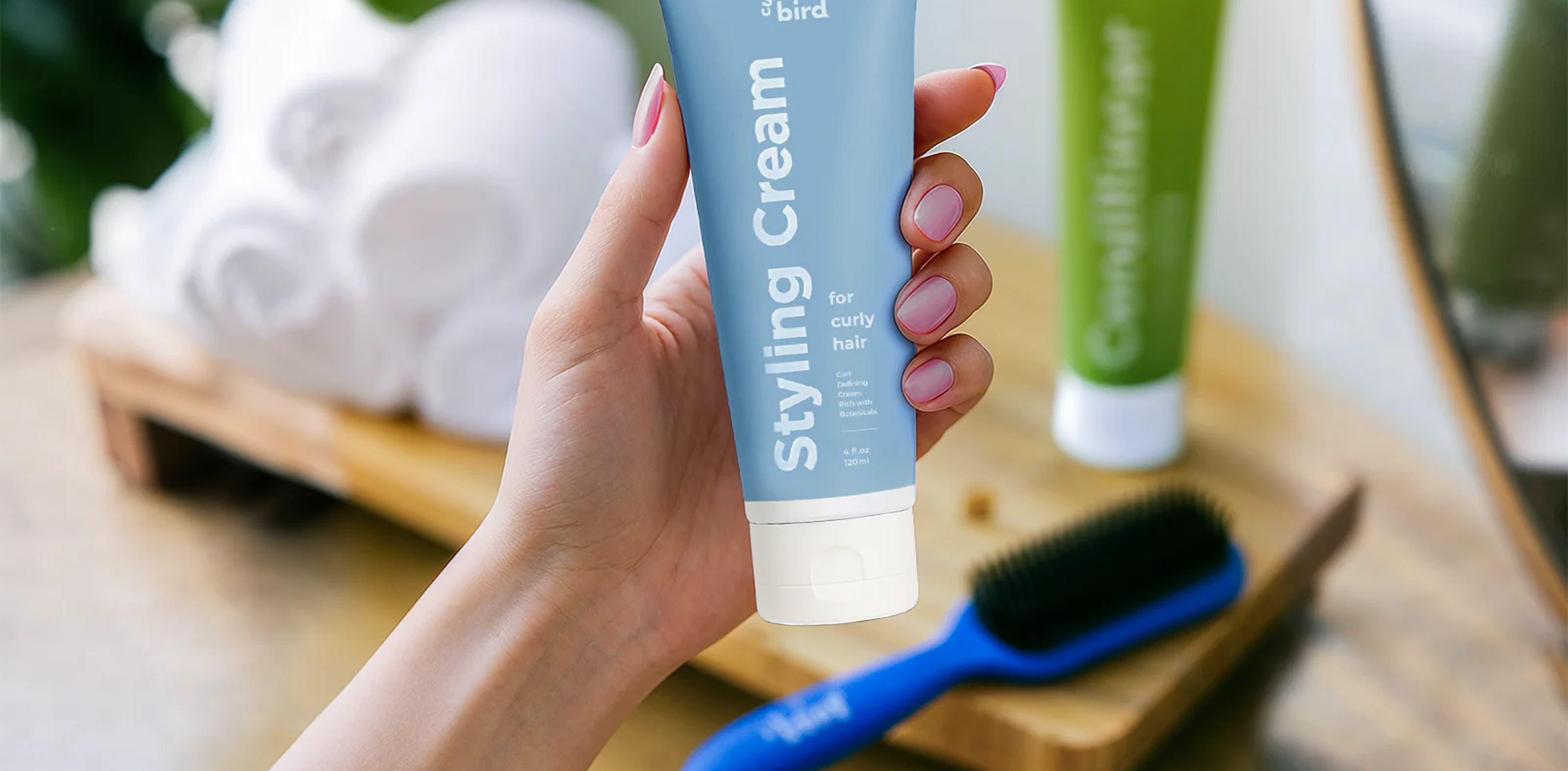A hand holds a light blue tube of Styling Cream with a hairbrush and towels in the background.