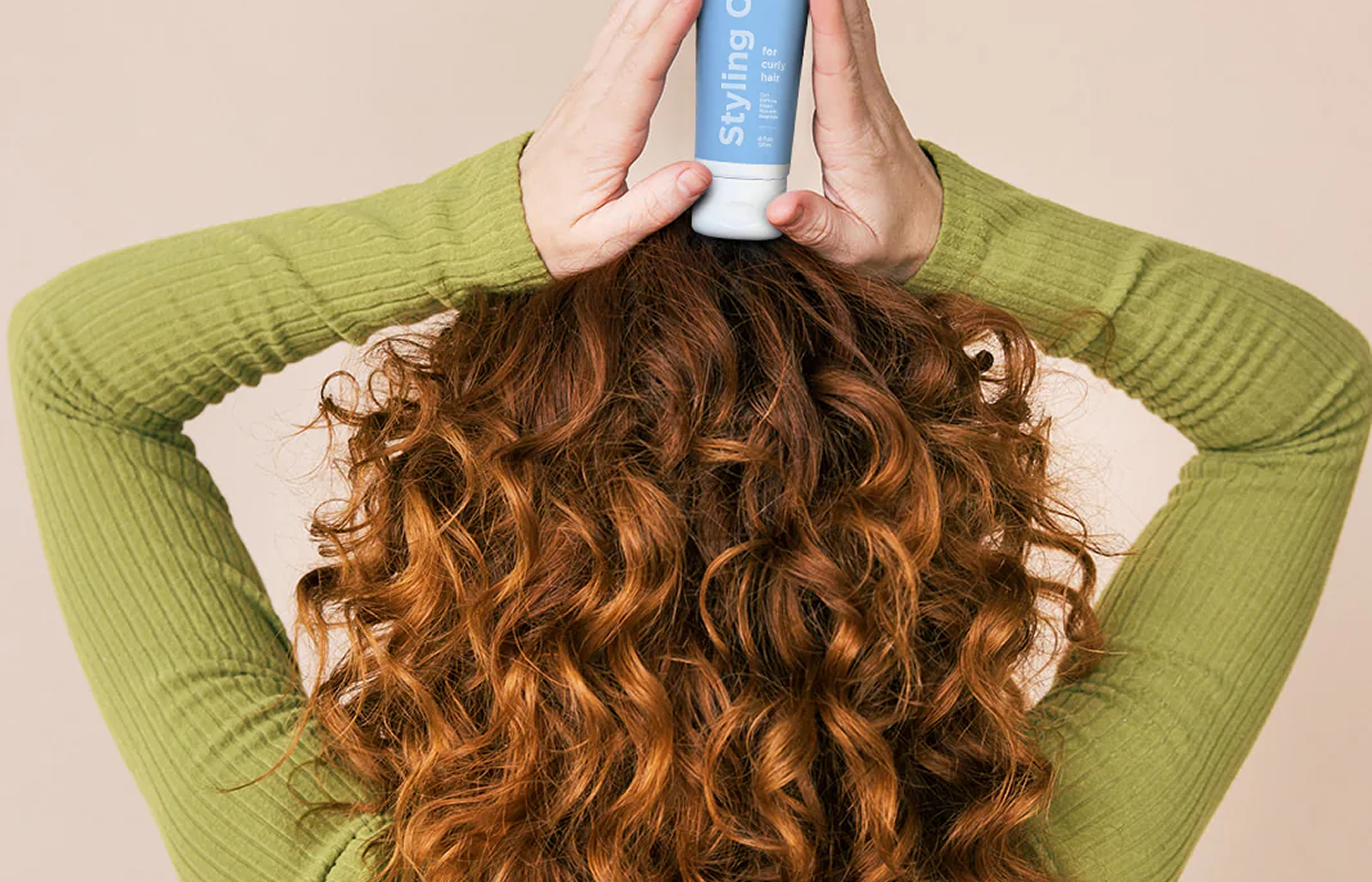 The back of a person with curly hair, holding a blue tube of styling product on their head.